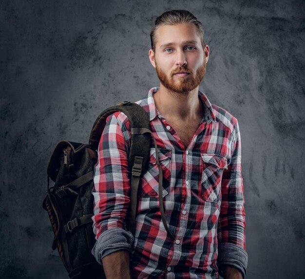 Stylish redhead urban traveller male holds backpack on grey background in a studio.