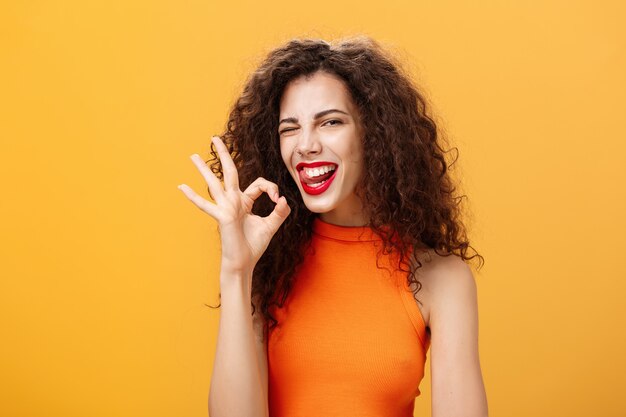 Stylish self-assured caucasian girlfriend with curly hairstyle winking joyfully and sticking out tongue showing perfect or okay gesture assuring she got everything under control over orange wall.