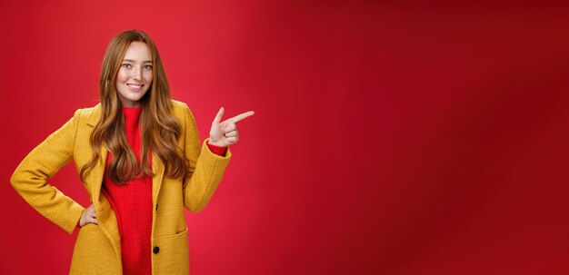 Stylish and selfassured attractive redhead woman with freckles in yellow outdoor coat pointing right