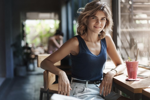 Stylish selfassured charming modern young woman sit trendy urban space cafe lean table bar near window smiling turn camera sassy drink healthy smoothie finish talking smartphone