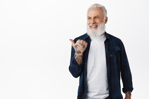 Stylish senior man, mature guy with beard pointing left, smiling and looking happy aside at copy space, showing advertisement, standing in modern hipster clothes against white background.