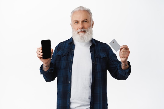 Stylish senior man with beard showing smartphone screen and credit card recommending shopping app or banking feature standing over white background