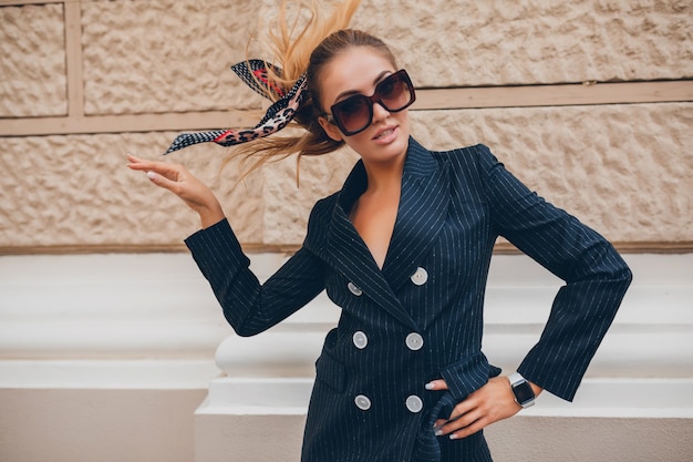 Stylish sexy woman dressed in elegant tuxedo suit walking in city on summer spring day