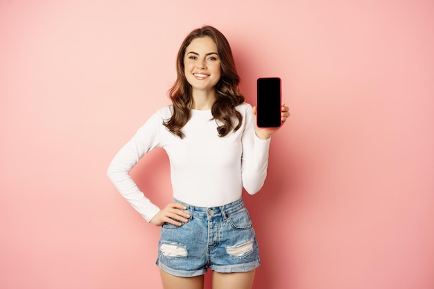 Stylish sexy woman showing mobile phone screen, smartphone app interface and smiling, demonstarting online shop, standing over pink background