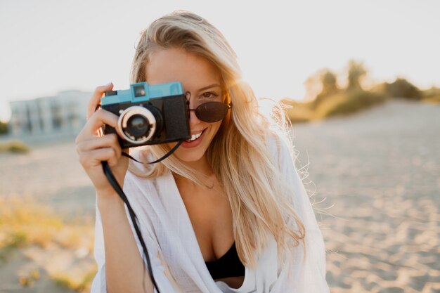써니 비치에서 포즈를 취하는 레트로 카메라로 세련 된 매끈한 소녀. 여름 방학. 열대 분위기. 자유와 여행 개념.