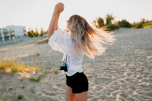 日当たりの良い夜のビーチでポーズをとってレトロなカメラでスタイリッシュな形の良い女の子。夏休み。トロピカルムード。自由と旅行のコンセプト。