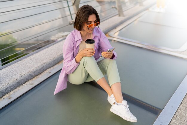 モダンな橋の上に座って、コーヒーを楽しんで、携帯電話を使用してスタイリッシュな短い髪の女性