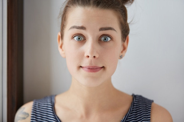 Free photo stylish shot of surprised scared caucasian girl with brown hair, charming blue eyes, white skin. young female with tattoo looks lovely in her natural amusement with closed mouth, raised eyebrows.