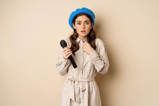 Stylish singer woman with microphone touching throat feeling ill standing over beige background