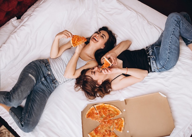 スタイリッシュな姉妹がピザをスライスしてベッドに横たわっている