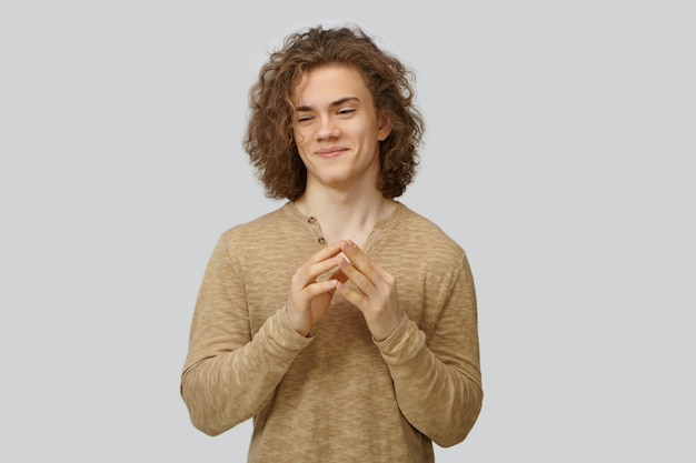 Free photo stylish sly young european man with voluminous curly hairstyle smiling mysteriously and clasping hands, scheming, planning tricky joke, going to prunk his best friend. human emotions