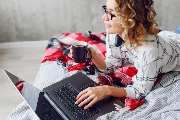 노트북을 사용하는 안경에 세련된 스마트 쾌활한 여자, 아늑한 침대에 누워, 커피를 마시는.
