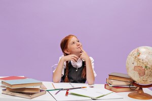 Stylish smart girl with red hair in school uniform is thoughtful and sitting at table with books notebooks and globe on lilac backdrop