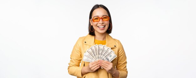 Stylish smiling asian girl holding money cash showing dollars and celebrating standing over white background