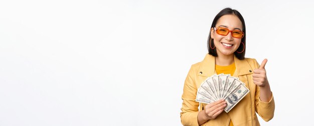 현금을 들고 달러를 보여주고 흰색 배경 위에 서 있는 것을 축하하는 세련된 미소 아시아 소녀