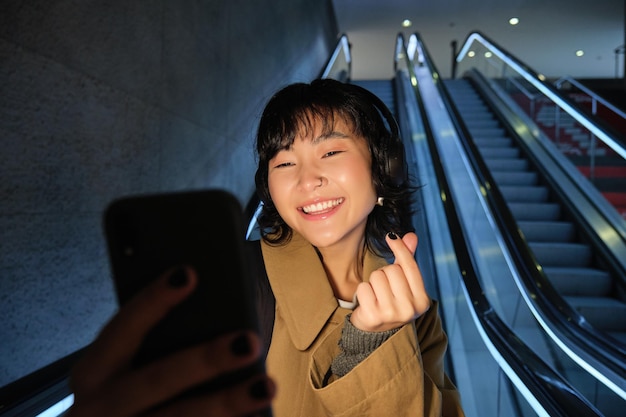 Free photo stylish smiling asian girl taking selfie on mobile phone while riding escalator going down to metro