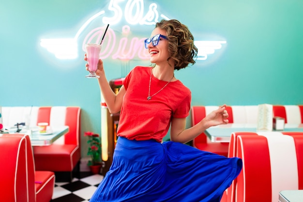 Stylish smiling woman in colorful outfit in retro vintage 50's cafe dancing drinking milk shake cocktail in blue skirt and red shirt wearing sunglasses having fun in cheerful mood