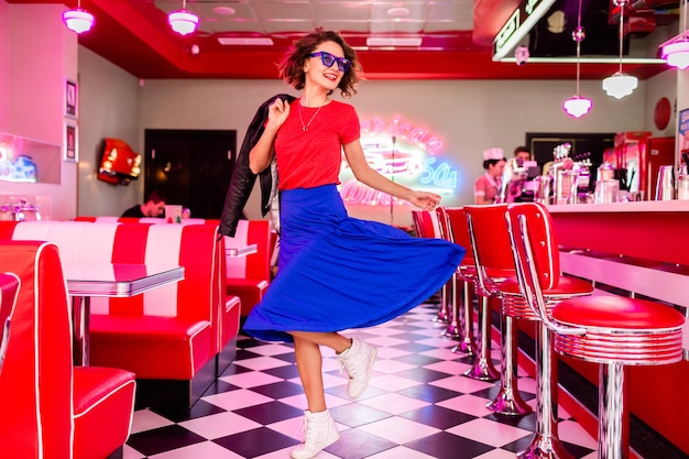 Stylish smiling woman in colorful outfit in retro vintage 50's cafe dancing wearing jacket, blue skirt and red shirt, sunglasses having fun in cheerful mood