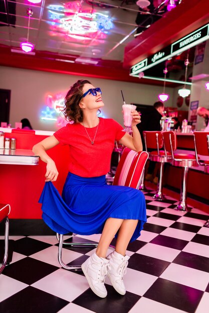 테이블에 앉아 복고풍 빈티지 50 년대 카페에서 화려한 옷을 입고 세련된 웃는 여자