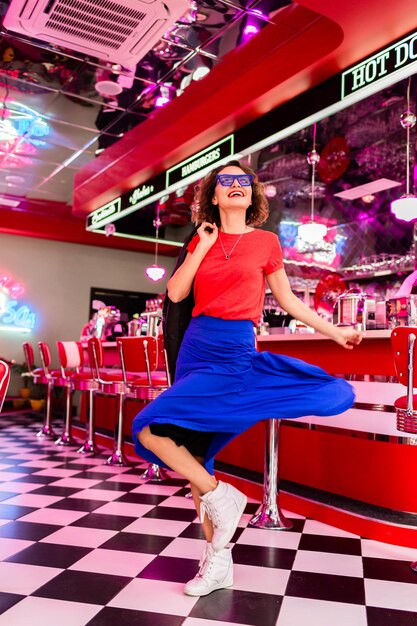 Stylish smiling woman in colorful outfit in retro vintage 50s cafe dancing wearing jacket blue skirt and red shirt sunglasses having fun in cheerful mood