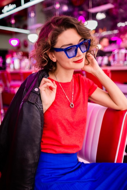 복고 빈티지 50년대 카페에서 화려한 옷을 입은 세련된 웃는 여성이 테이블에 앉아 재킷 파란색 치마와 빨간색 셔츠 선글라스를 착용하고 쾌활한 분위기에서 즐거운 시간을 보내고 있습니다.