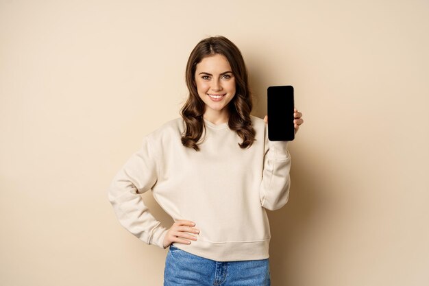 Stylish smiling woman showing smartphone screen mobile app interface standing over beige background