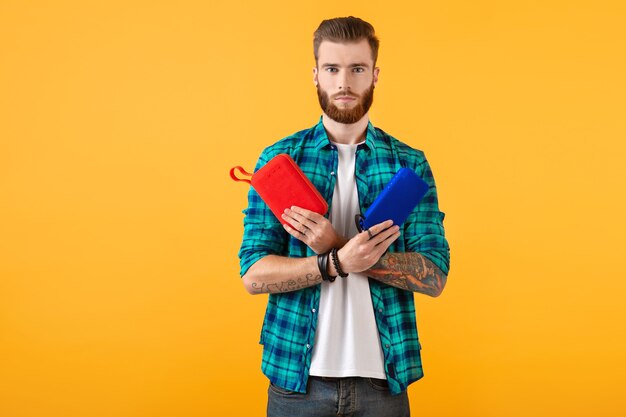 Stylish smiling young man holding wireless speaker happy listening to music colorful style happy mood isolated on yellow wall