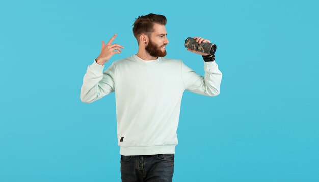 Stylish smiling young man holding wireless speaker listening to music on blue