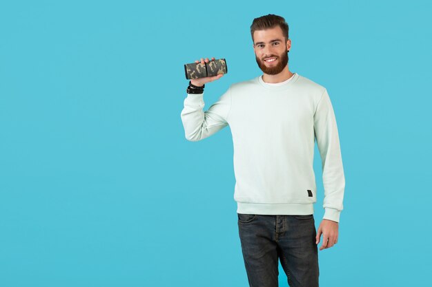 Stylish smiling young man holding wireless speaker listening to music colorful style happy mood isolated on blue wall