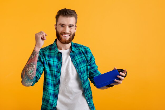 Stylish smiling young man holding wireless speaker listening to music on orange