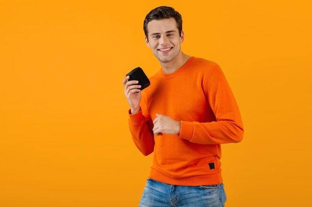 Stylish smiling young man in orange sweater holding wireless speaker happy listening to music having fun colorful style happy mood isolated on yellow background