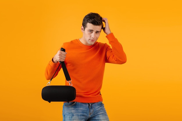 Stylish smiling young man in orange sweater holding wireless speaker happy listening to music having fun colorful style happy mood isolated on yellow background