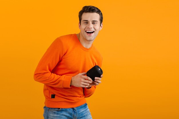 Stylish smiling young man in orange sweater holding wireless speaker happy listening to music having fun on orange