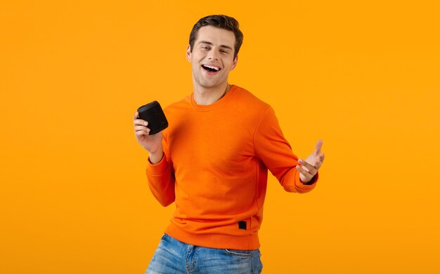 Stylish smiling young man in orange sweater holding wireless speaker happy listening to music having fun on orange