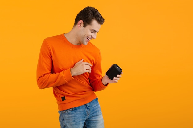 オレンジ色のセーターでスタイリッシュな笑顔の若い男は、オレンジ色で楽しんで音楽を聴いて幸せなワイヤレススピーカーを保持