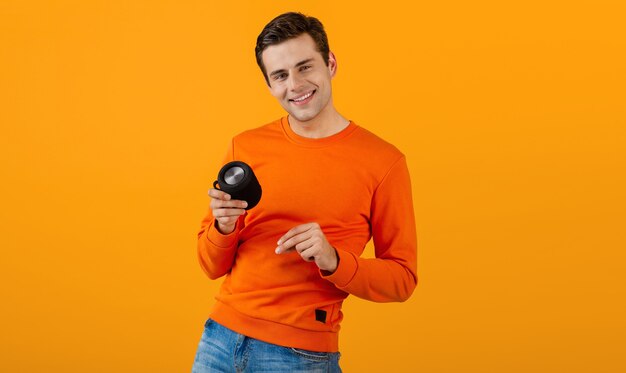 Stylish smiling young man in orange sweater holding wireless speaker happy listening to music having fun on orange