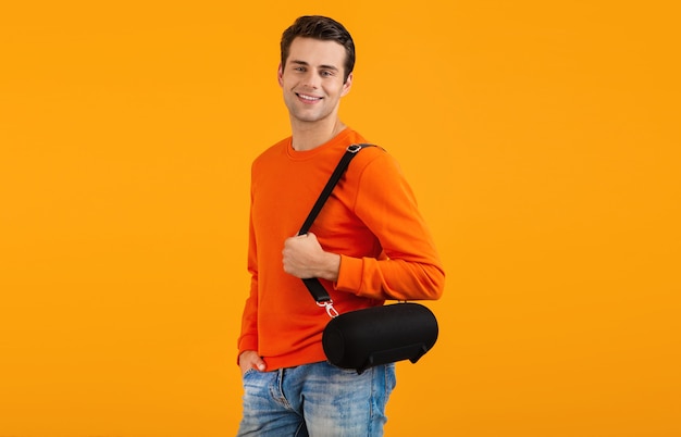 Stylish smiling young man in orange sweater holding wireless speaker happy listening to music having fun on orange