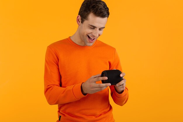 Stylish smiling young man in orange sweater holding wireless speaker happy listening to music having fun
