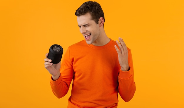 stylish smiling young man in orange sweater holding wireless speaker happy listening to music having fun