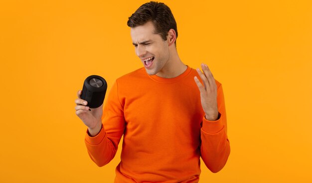 ワイヤレス スピーカーを保持しているオレンジ色のセーターを着たスタイリッシュな笑顔の若い男が楽しんで音楽を聴いて幸せ
