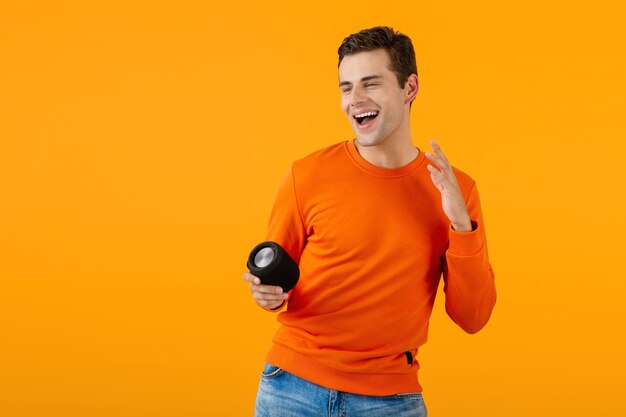 Стильный улыбающийся молодой человек в оранжевом свитере, держащий беспроводную колонку, счастлив, слушая музыку, весело проводя время