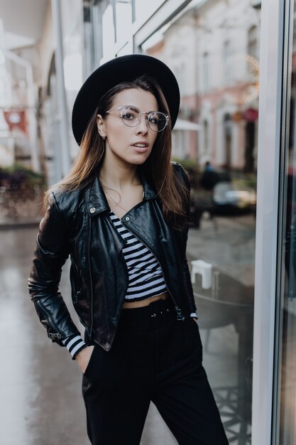 Stylish student girl walks in the city after rain near brilliant shop windows in cold weather in the day