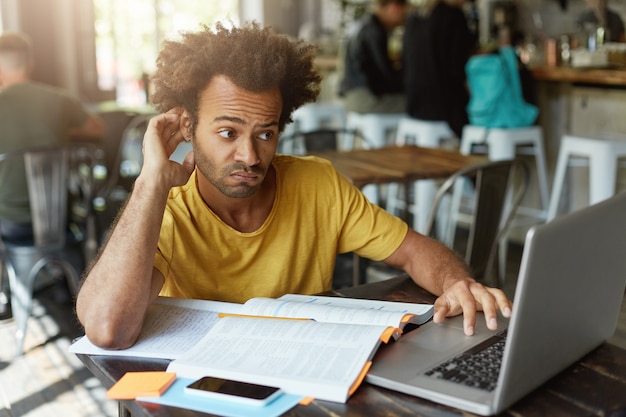 Stylish student with African hairstyle having doubtful look while looking at laptop computer not understanding new material trying to find good explanation in internet