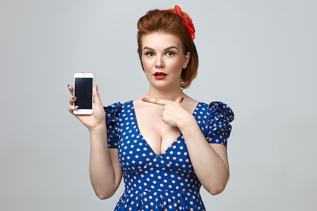 Stylish surprised young Caucasian lady in cut out dotted dress holding smrt phone, pointing finger at blank copy space screen