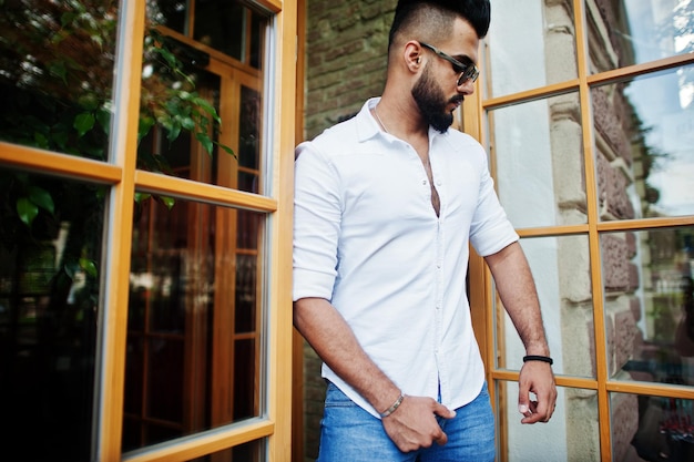 Stylish tall arabian man model in white shirt jeans and sunglasses posed at street of city Beard attractive arab guy against entrance door of restaurant