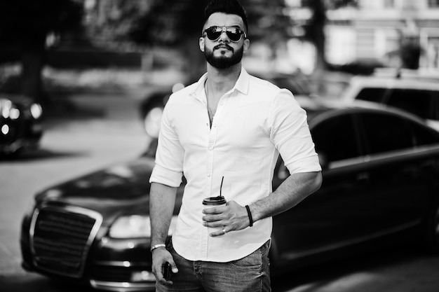 Стильный высокий арабский мужчина в белых джинсах и солнцезащитных очках позирует на улице города Борода привлекательный арабский парень с чашкой кофе на фоне черной деловой машины