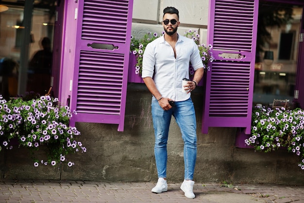 흰색 셔츠 청바지와 선글라스를 쓴 세련된 키 큰 아라비아 남자 모델은 보라색 창문에 커피 한 잔을 들고 도시 수염 매력적인 아랍 남자의 거리에서 포즈를 취했습니다.