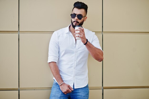 Stylish tall arabian man model in white shirt jeans and sunglasses posed at street of city Beard attractive arab guy with cup drinking coffee against golden wall