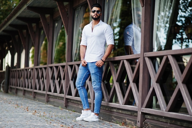 흰색 셔츠 청바지와 선글라스를 입은 세련된 키 큰 아라비아 남자 모델은 도시의 거리에서 포즈를 취했습니다. 수염 매력적인 아랍 남자