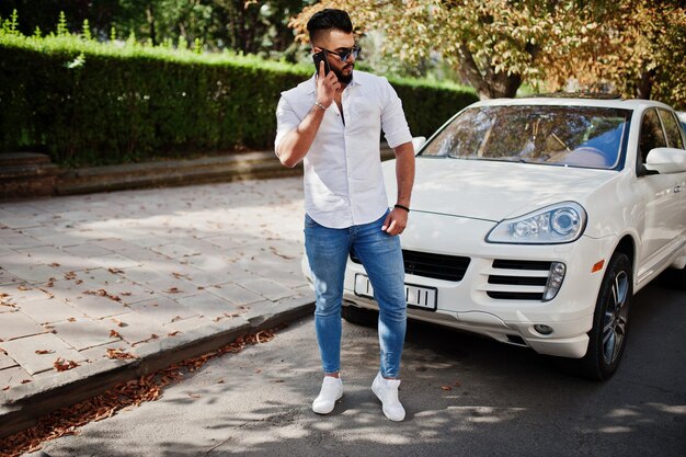 Stylish tall arabian man model in white shirt jeans and sunglasses posed at street of city Beard rich attractive arab guy against white suv car speaking on mobile phone
